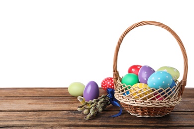 Photo of Colorful Easter eggs in wicker basket and willow branches on wooden table against white background. Space for text