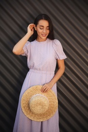 Beautiful young woman in stylish violet dress and straw hat near dark corrugated metal wall