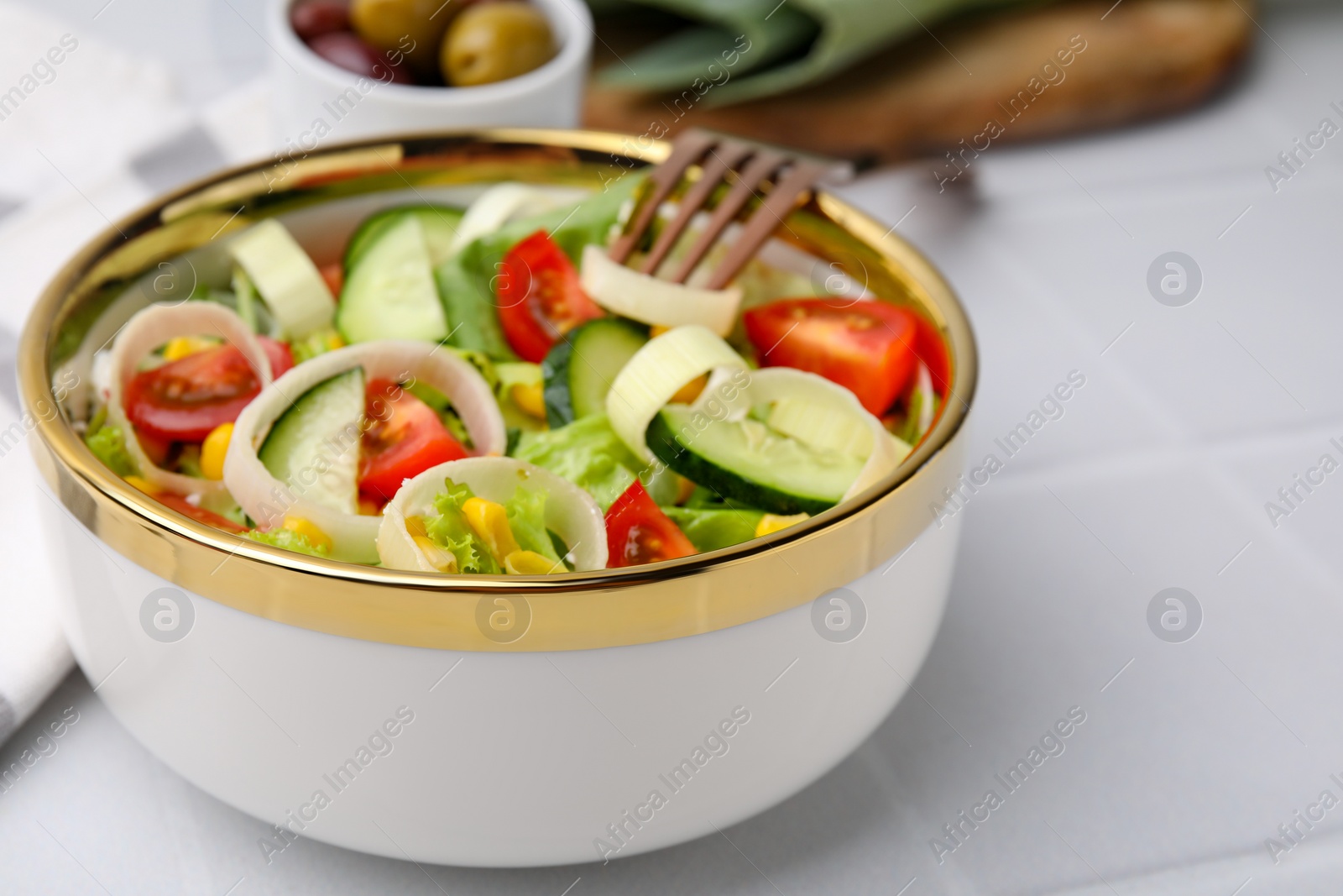 Photo of Bowl of tasty salad with leek, tomatoes and cucumbers on white tiled table, closeup