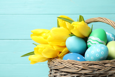 Photo of Easter eggs and yellow tulips in wicker basket on light blue background, closeup