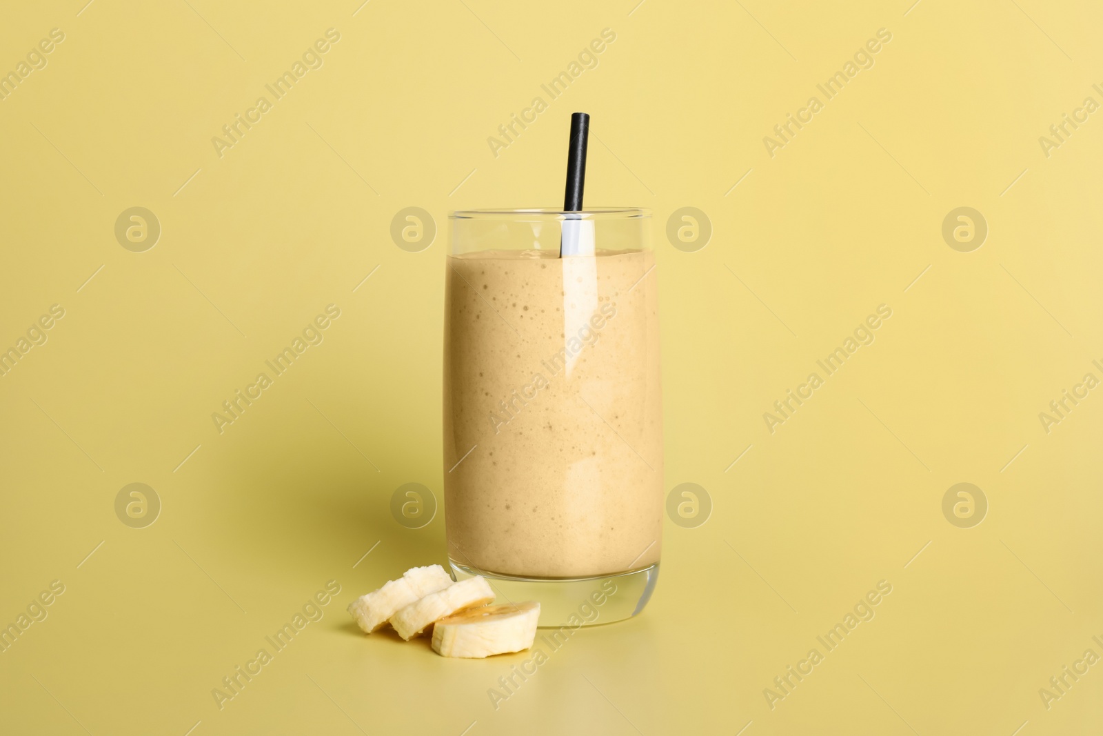 Photo of Glass of tasty smoothie with straw and cut banana on pale yellow background