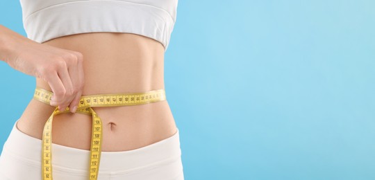 Slim woman measuring waist with tape on light blue background, closeup and space for text. Weight loss