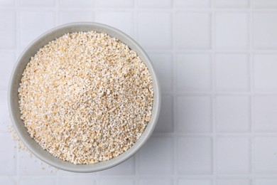 Photo of Dry barley groats in bowl on white tiled table, top view. Space for text