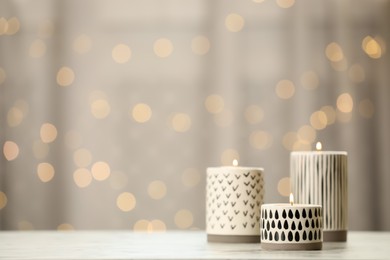 Photo of Burning candles in stylish holders on white table against blurred festive lights, space for text