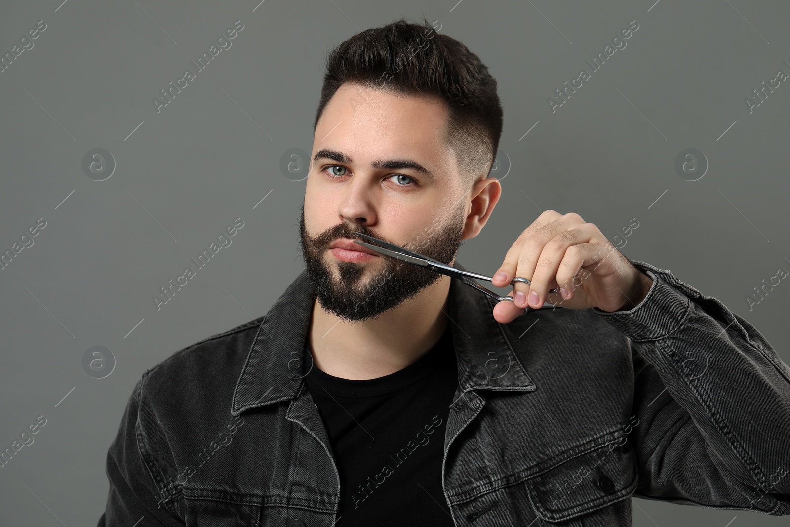 Photo of Handsome young man trimming mustache with scissors on grey background