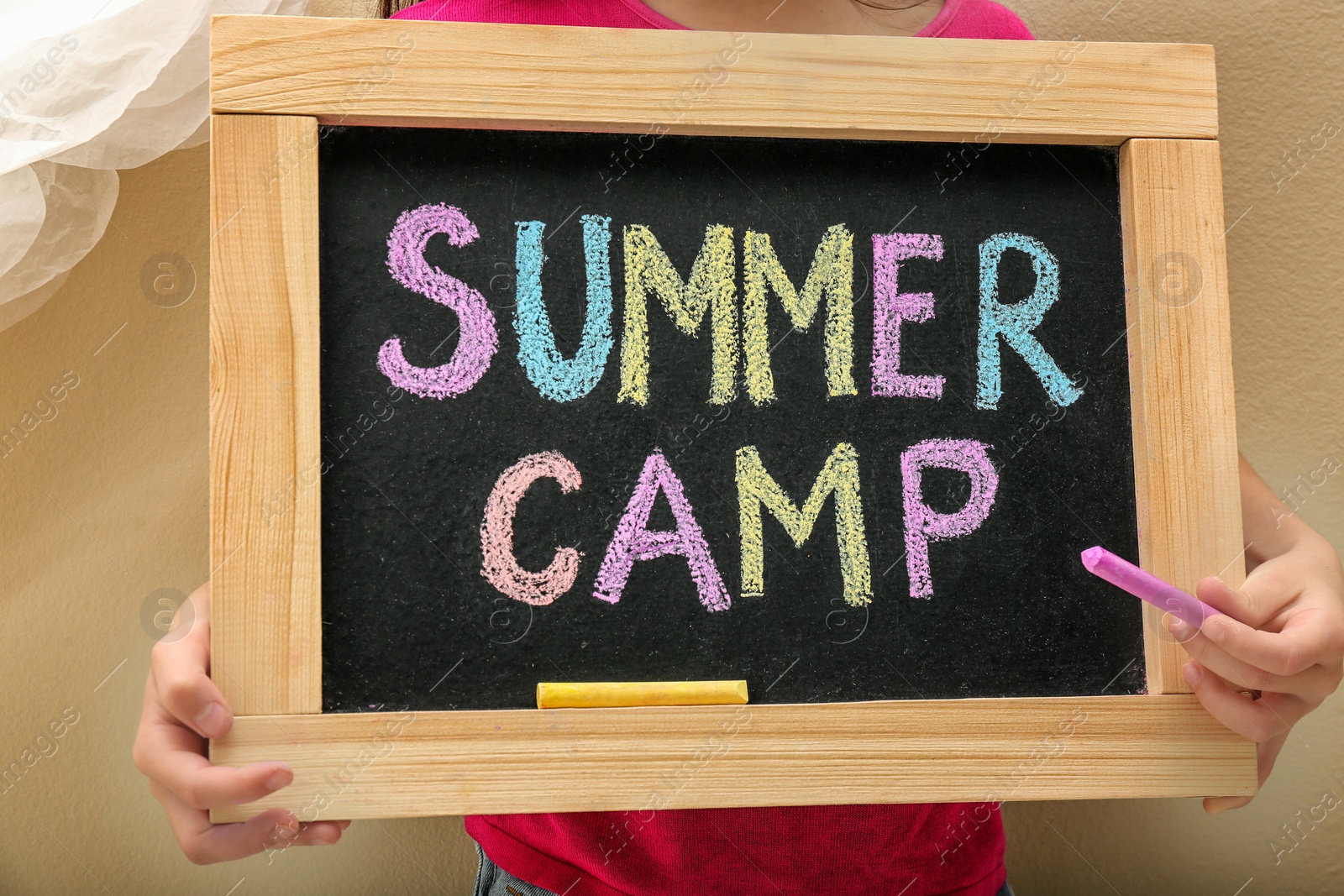 Photo of Child writing SUMMER CAMP with colorful chalk on blackboard, closeup