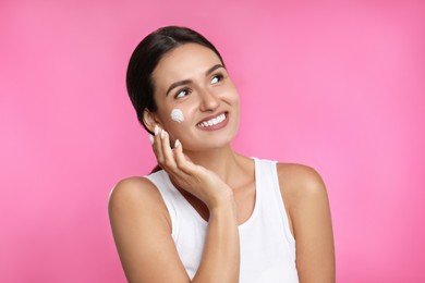 Young woman applying facial cream on pink background