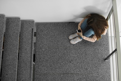 Photo of Upset teenage girl with smartphone sitting on staircase indoors, above view