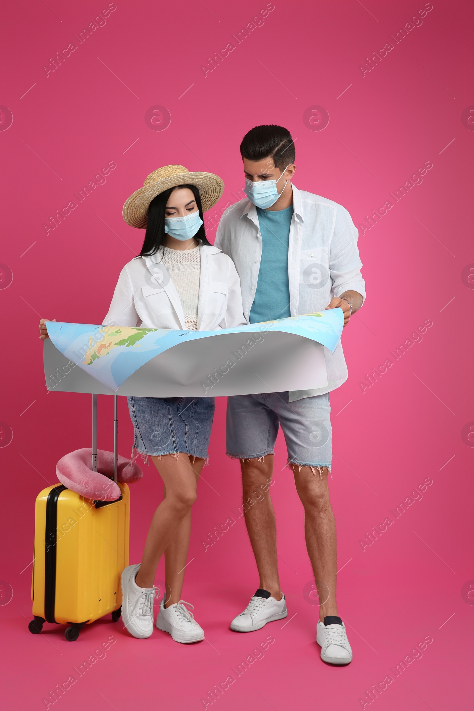 Photo of Couple of tourists in medical masks with map and suitcase on pink background. Travelling during coronavirus pandemic