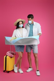 Couple of tourists in medical masks with map and suitcase on pink background. Travelling during coronavirus pandemic