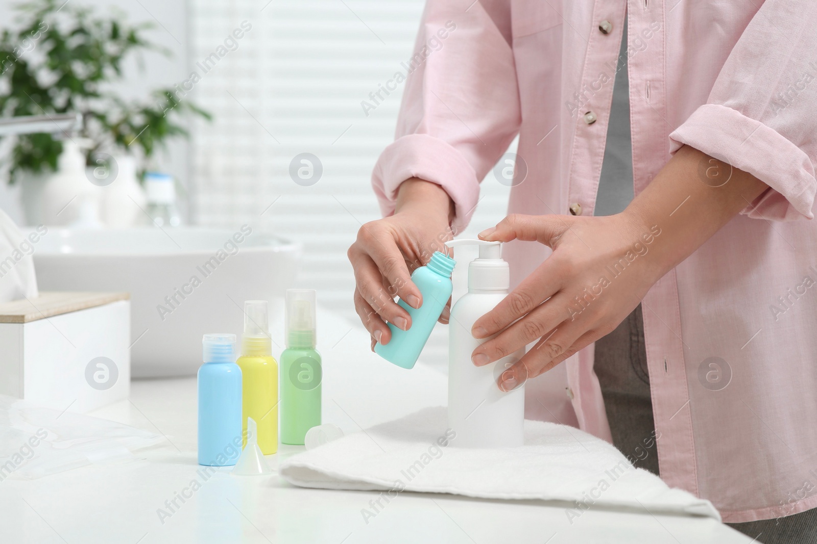 Photo of Woman pouring cosmetic product into plastic bottle at white countertop in bathroom, closeup. Bath accessories