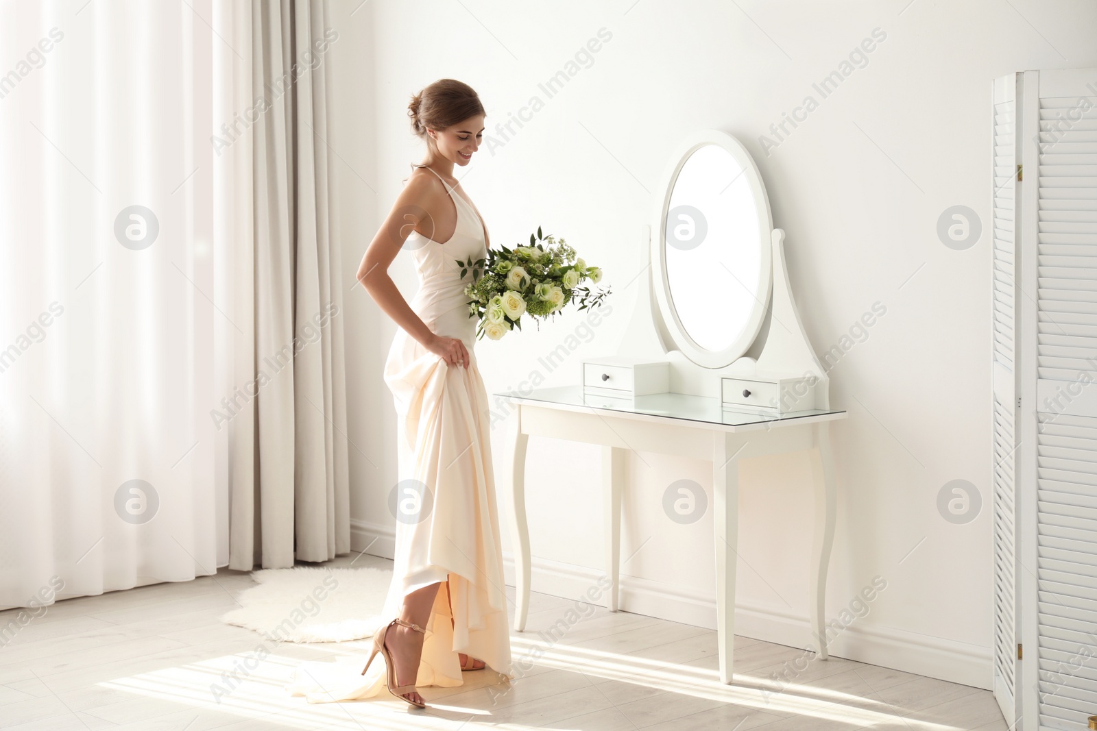 Photo of Young bride in wedding dress with beautiful bouquet near mirror indoors