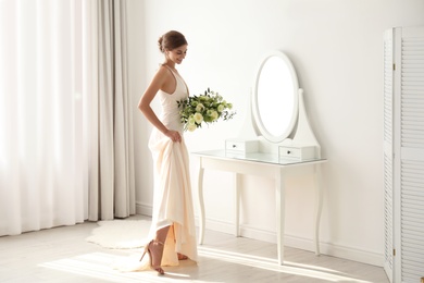 Young bride in wedding dress with beautiful bouquet near mirror indoors