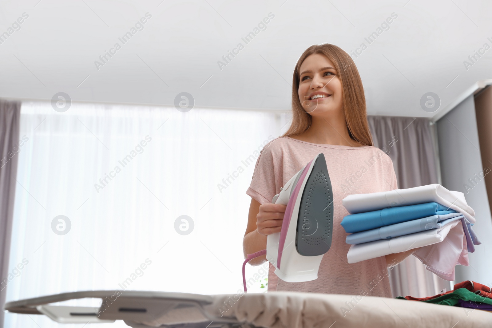 Photo of Young woman with folded clothes and iron at home. Space for text