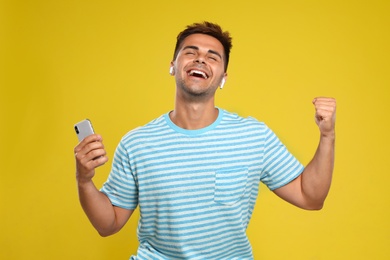 Happy young man with smartphone listening to music through wireless earphones on yellow background