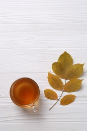 Small branch with autumn leaves and cup of delicious tea on white wooden table, flat lay