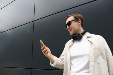 Handsome young man with headphones and modern smartphone near black wall, space for text