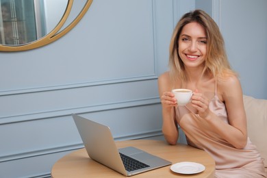 Photo of Happy young woman with cup of coffee and laptop at table indoors in morning