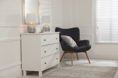 White chest of drawers, armchair and mirror in room. Interior design