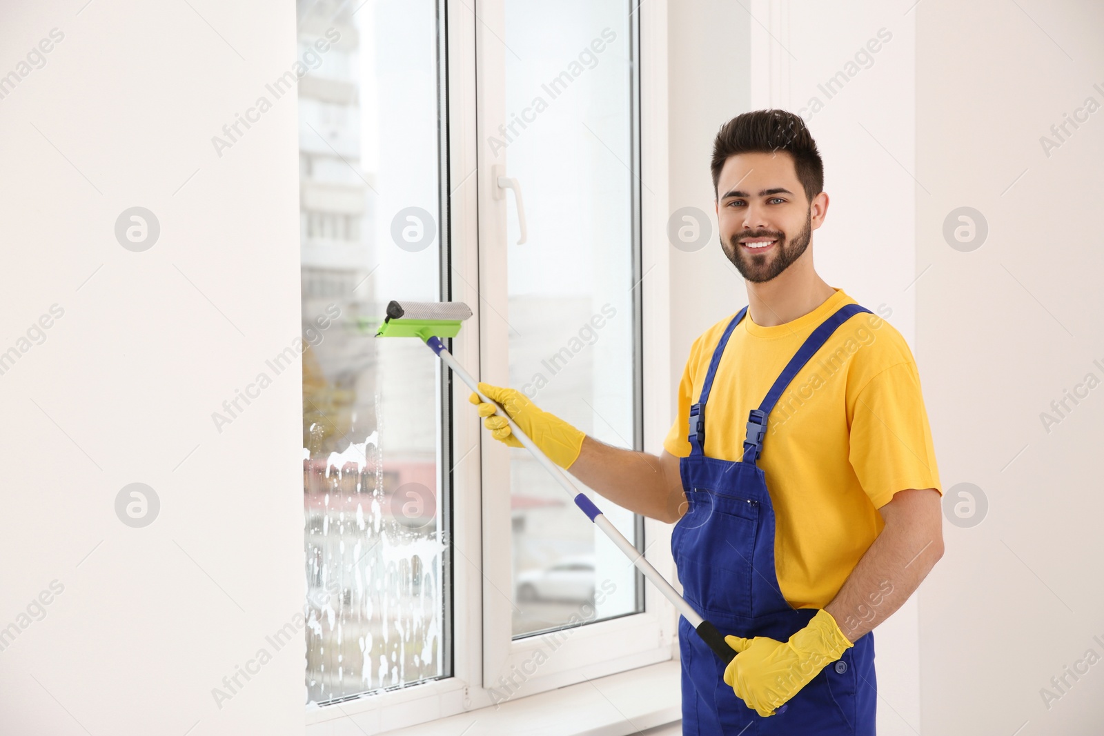 Photo of Professional young janitor in uniform cleaning window indoors