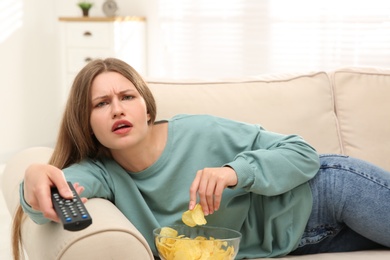 Lazy young woman with bowl of chips watching TV on sofa at home