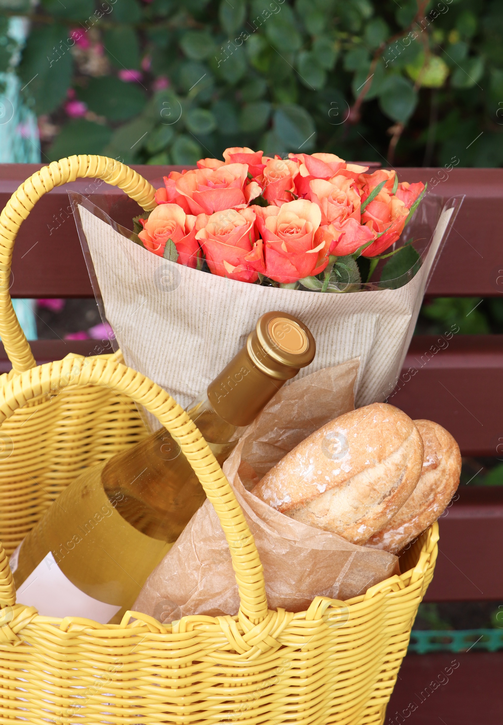 Photo of Beautiful roses, bottle of wine and baguettes in yellow wicker bag on bench outdoors