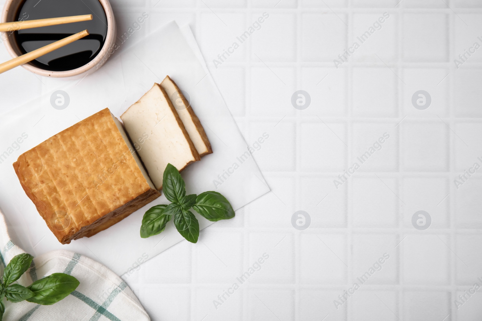 Photo of Delicious smoked tofu, soy sauce and basil on tiled table, flat lay. Space for text