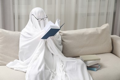 Photo of Creepy ghost. Person covered with white sheet reading book on sofa at home