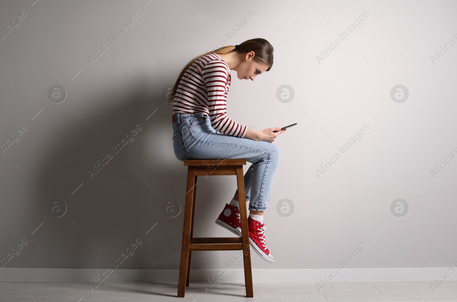 Photo of Woman with bad posture using tablet while sitting on stool near light grey wall indoors