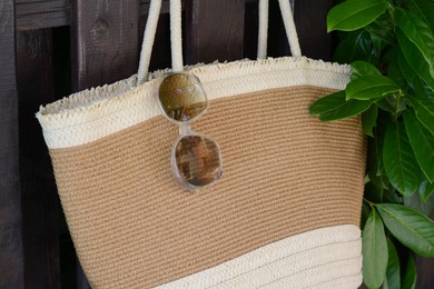 Photo of Stylish bag with sunglasses hanging on wooden fence outdoors, closeup. Beach accessories