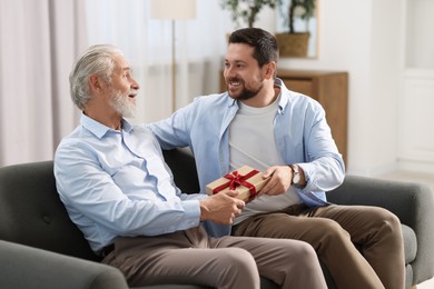 Photo of Son giving gift box to his dad on sofa at home