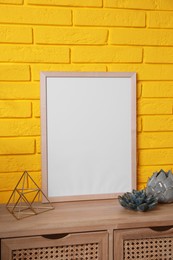 Empty frame and decor on wooden cabinet near yellow brick wall. Mockup for design