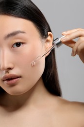 Beautiful young woman applying cosmetic serum onto her face on grey background