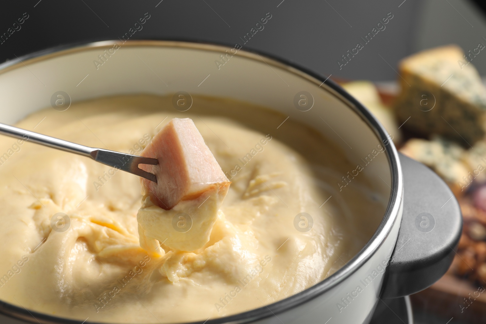 Photo of Dipping piece of ham into fondue pot with melted cheese on grey background, closeup