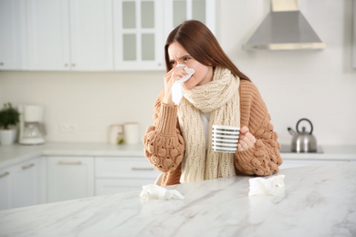 Sick young woman with cup of hot drink and tissues in kitchen. Influenza virus