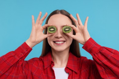 Young woman holding halves of kiwi near her eyes on light blue background