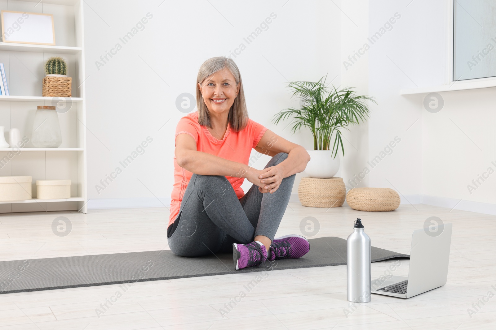 Photo of Senior woman in sportswear sitting on fitness mat near laptop at home