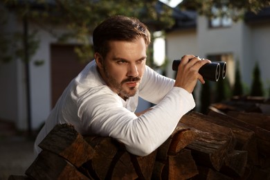 Photo of Concept of private life. Curious man with binoculars spying on neighbours over firewood outdoors