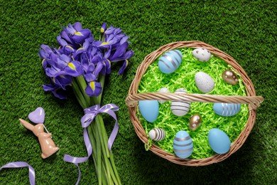 Photo of Easter basket with painted eggs, iris flowers and figure of rabbit on green grass, flat lay