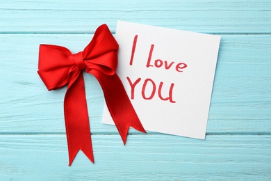 Photo of Red bow and note with words I LOVE YOU on blue wooden table