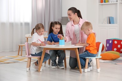 Photo of Nursery teacher and group of cute little children drawing at desk in kindergarten. Playtime activities