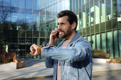 Photo of Emotional man talking on smartphone and looking at watch near building. Being late concept