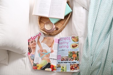 Different lifestyle magazines, books and coffee on bed, above view