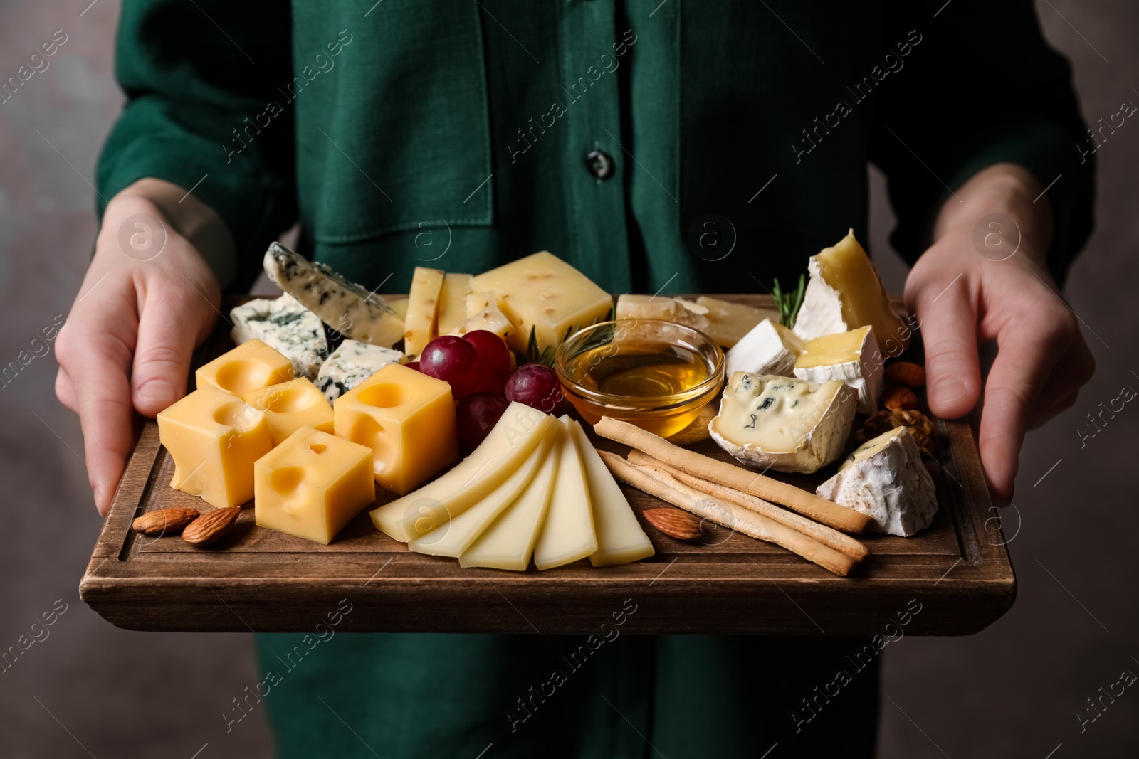 Photo of Woman holding cheese plate with honey, grissini and grapes on grey background, closeup