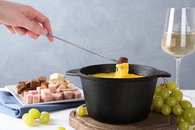 Photo of Woman dipping piece of bread into fondue pot with tasty melted cheese at white table against gray background, closeup