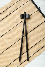 Photo of Bamboo mat with pair of black chopsticks and rest on white table, top view