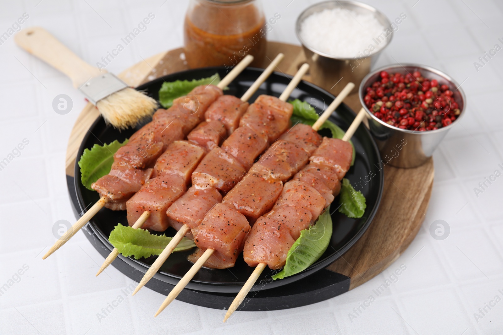 Photo of Wooden skewers with cut raw marinated meat, spices and basting brush on white tiled table