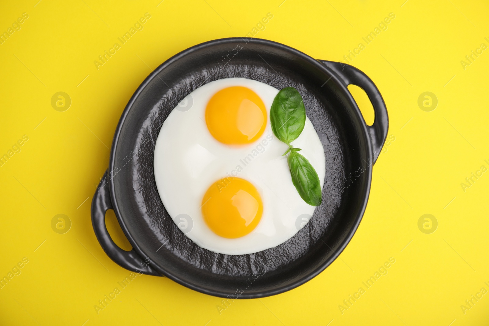 Photo of Tasty fried eggs with basil in pan on yellow background, top view