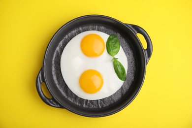 Tasty fried eggs with basil in pan on yellow background, top view