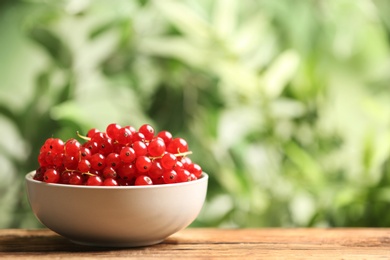 Photo of Ripe red currants in bowl on wooden table against blurred background. Space for text